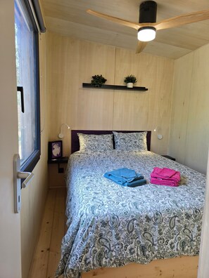 Un dormitorio con ventilador de techo de madera, cama con ropa de cama estampada, mesita de noche con lámpara y ventana con cortinas.