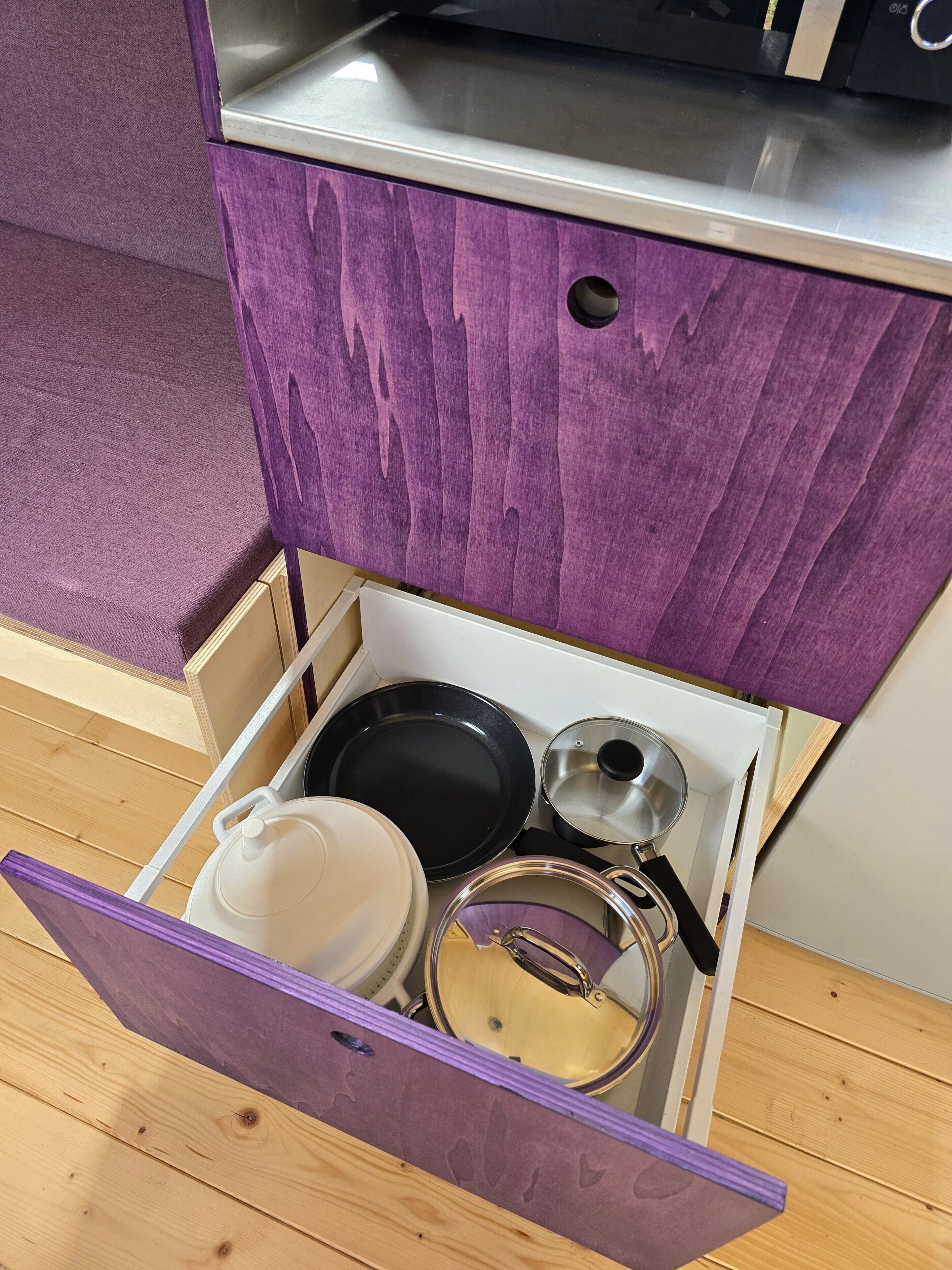 Un tiroir de cuisine dont l’intérieur est violet, contenant une casserole blanche, une poêle noire et une casserole en acier inoxydable avec un couvercle.