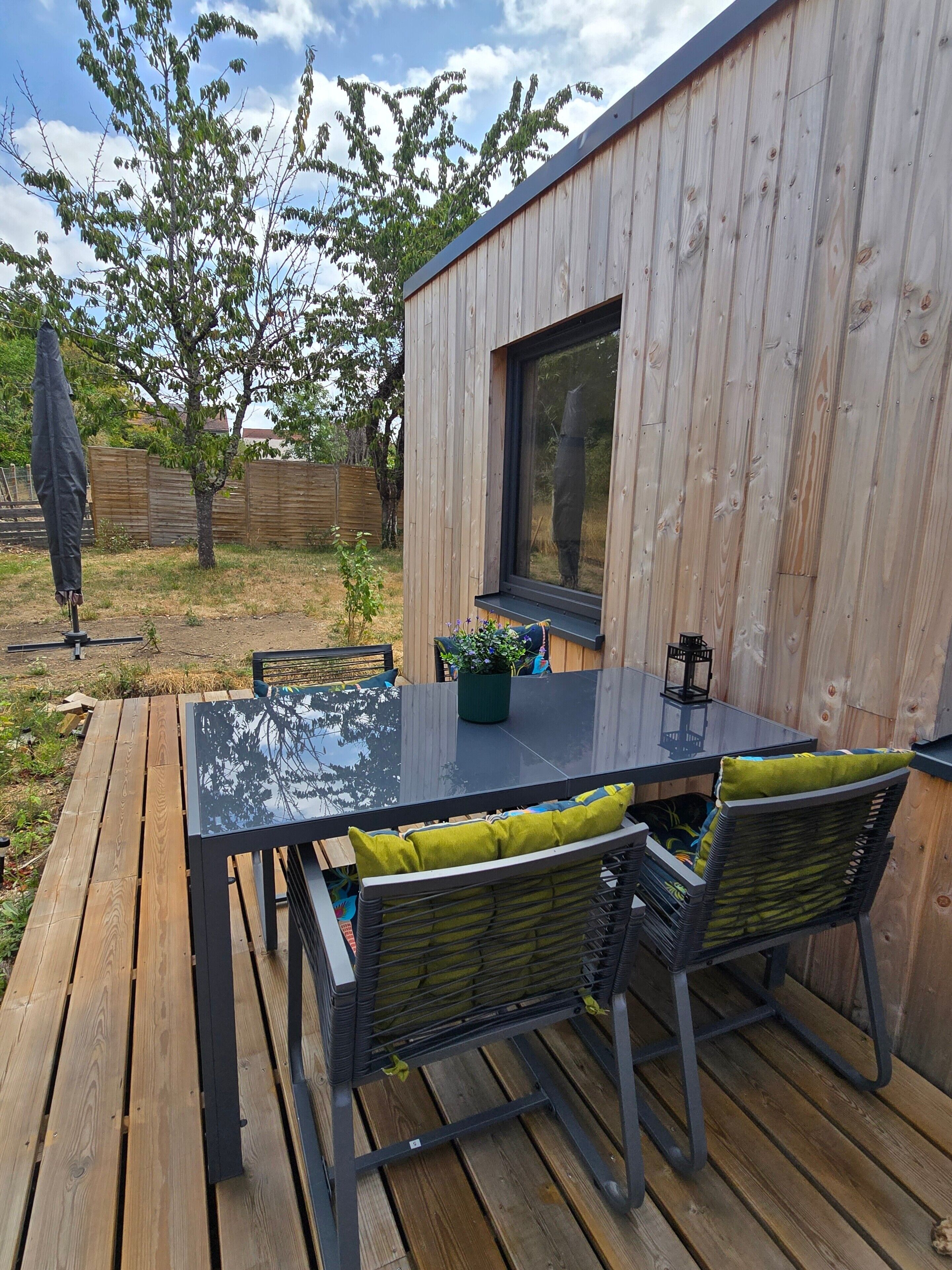 An outdoor dining area with a glass-top table and metal chairs, set against a wooden wall with a window.