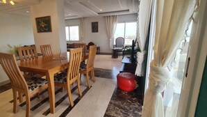 Ein Essbereich mit einem Holztisch und Stühlen, einem roten Pouf und Blick ins Freie durch ein Fenster.