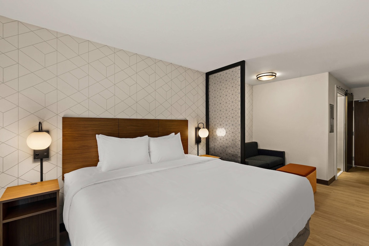Una camera d'hotel moderna con un letto grande, due comodini, un tavolino e una lampada a parete, oltre a un pannello decorativo con motivo geometrico.