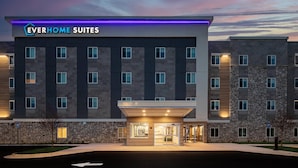 Un moderno edificio de hotel con un letrero que dice 'Everhome Suites'. Cuenta con fachada de piedra, múltiples ventanas y una entrada bien iluminada.