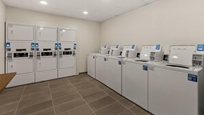 Una sala de lavandería con varias lavadoras y secadoras, piso de baldosas y techo blanco con iluminación empotrada.