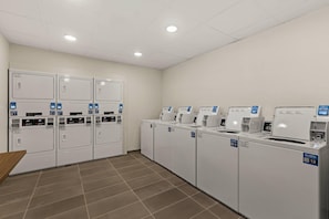 Lavanderia dotata di diverse lavatrici e asciugatrici, pavimento piastrellato e soffitto bianco con faretti.