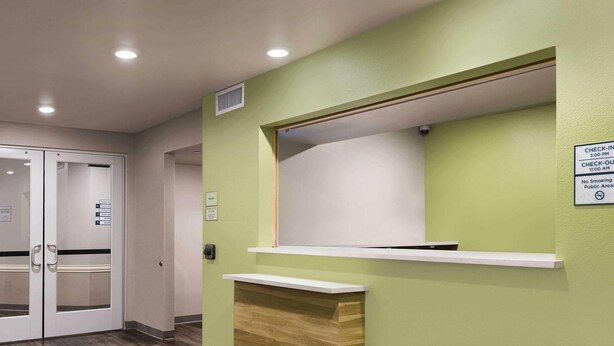 A reception desk with a green wall, a wooden counter, and a sign indicating check-in/out times. - Woodspring Suites Rock Hill Charlotte (Rock Hill)