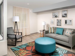 A modern living room with a grey sofa, a blue ottoman, a black chair, a floor lamp, and a patterned rug.