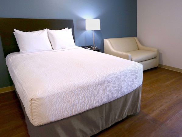 A neatly made bed with white linens, a bedside lamp, and a chair in a room with wooden flooring and a blue wall.