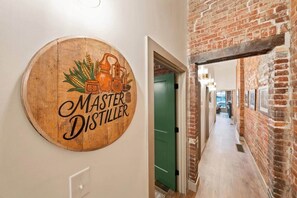 Ein hölzernes Schild mit der Aufschrift 'Master Distiller' und landwirtschaftlichen Illustrationen, eine grüne Tür und ein Korridor mit Backsteinwand.