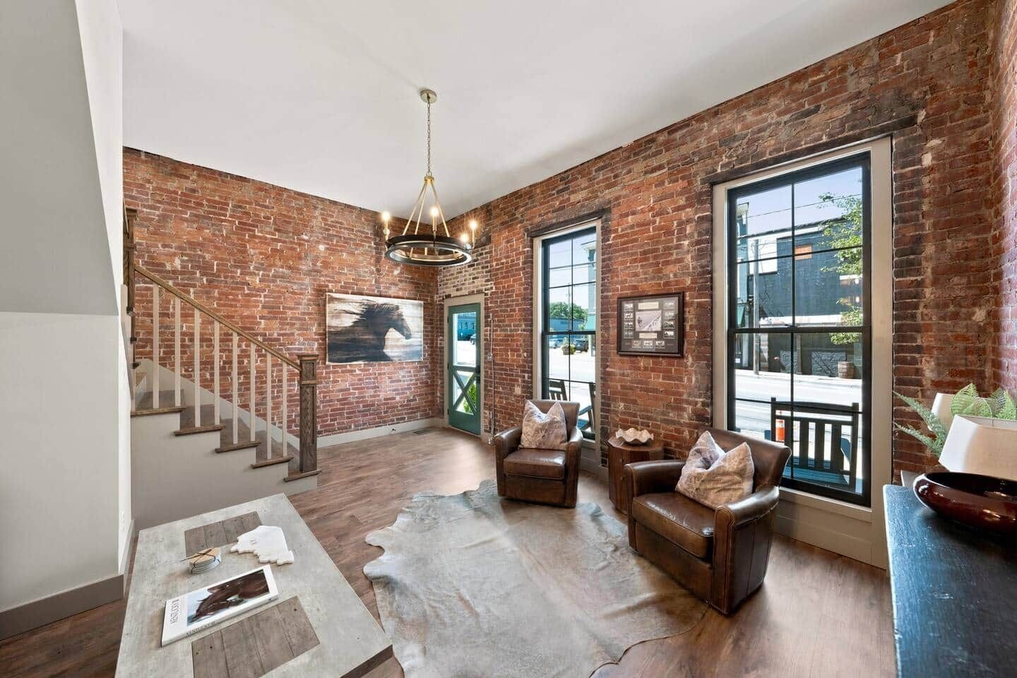 A spacious living area with a brick wall, a staircase, leather sofas, and a chandelier.