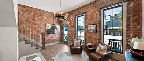 A spacious living area with a brick wall, a staircase, leather sofas, and a chandelier.