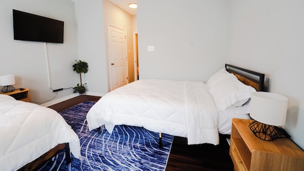 A hotel room with two beds, a flat-screen TV mounted on the wall, a bedside table with a lamp, and a door leading to another room.