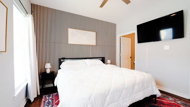 A hotel room with a bed, a television mounted on the wall, a patterned wall, and a window with curtains.