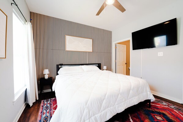 A hotel room with a bed, a television mounted on the wall, a patterned wall, and a window with curtains.