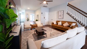A modern living room with a brown leather sofa, a white sofa, a coffee table, and a patterned rug. There is a staircase with a wooden railing and a ceiling fan.