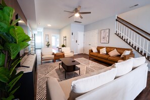 A modern living room with a brown leather sofa, a white sofa, a coffee table, and a patterned rug. There is a staircase with a wooden railing and a ceiling fan.