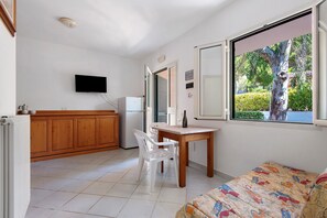 A compact room with a kitchenette, a dining area with a table and chairs, a bed with a colorful bedspread, and a window with a view of greenery. - Two-room Apartment with Veranda and Garden 100m from the Sea - Villaggio Sole e Mare (Rio)