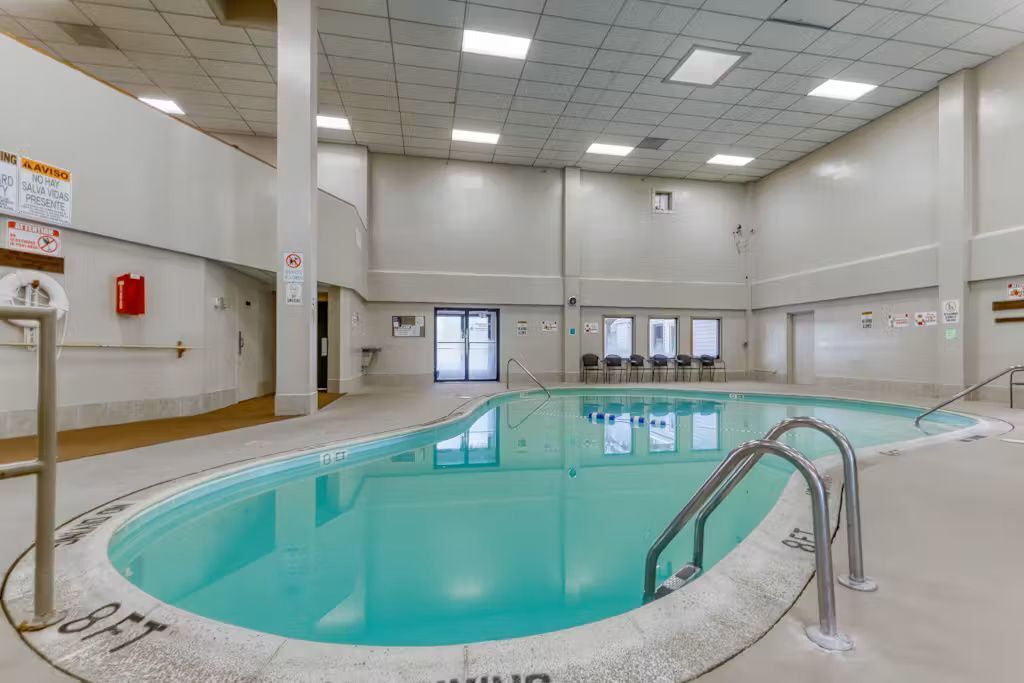 An indoor swimming pool with clear water, surrounded by a tiled floor and walls, and equipped with safety railings.