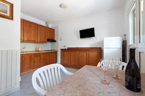 A kitchen with wooden cabinets, a TV mounted on the wall, a refrigerator, and a dining table with two wine glasses and a bottle. - Two-room Apartment with Veranda and Garden 100m from the Sea - Villaggio Sole e Mare (Rio)