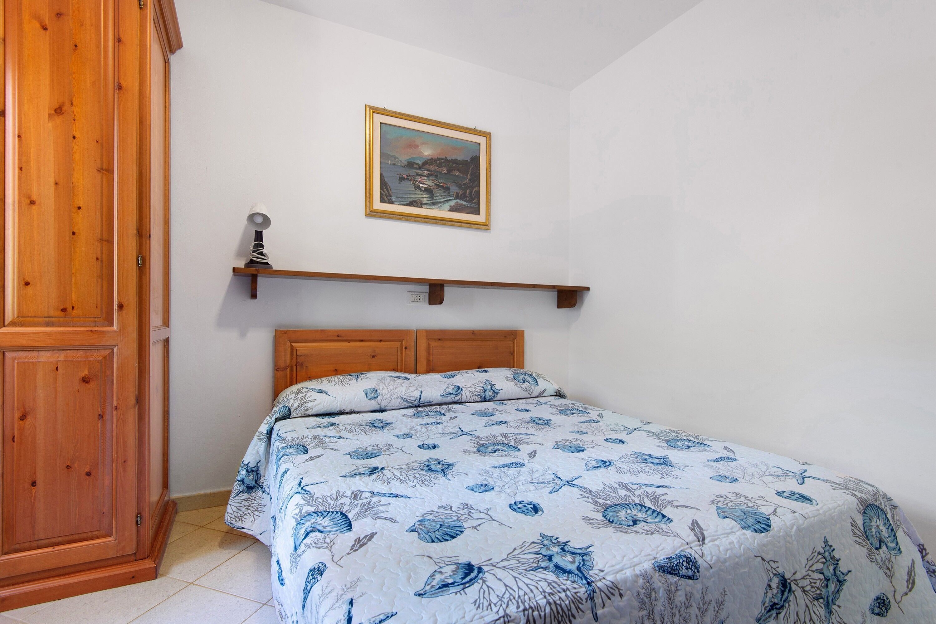 Une chambre à coucher avec une tête de lit en bois, un lit recouvert d’une couvre-lit à motifs floraux bleus, une étagère en bois ornée d’un tableau encadré et une armoire en bois.