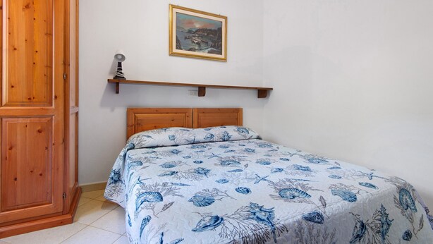 A bedroom with a wooden bed, a nightstand, a painting on the wall, and a wooden wardrobe. - Two-room Apartment with Veranda and Garden 100m from the Sea - Villaggio Sole e Mare (Rio)