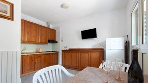 A kitchen with wooden cabinets, a TV mounted on the wall, a refrigerator, and a dining table with two wine glasses and a bottle. - Two-room Apartment with Veranda and Garden 100m from the Sea - Villaggio Sole e Mare (Rio)