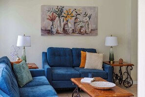 Ein Wohnzimmer mit blauem Sofa, zwei Nachttische mit Lampen, einem Couchtisch mit Buch und dekorativem Teller, und einem Wandbild mit Vasen.