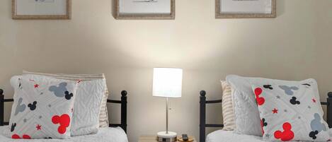 A hotel room with two beds, each with Mickey Mouse themed pillows, a bedside table with a lamp, and framed pictures on the wall.