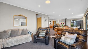 Ein Wohnzimmer mit einer Couch, zwei Sesseln, einem Esstisch mit Stühlen, einer Küche im Hintergrund und einem großen Fenster.