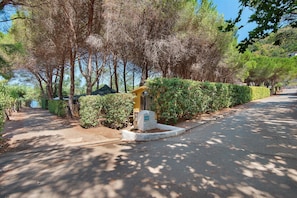 A paved pathway flanked by neatly trimmed hedges and trees, leading to a building with a visible entrance. - Two-room Apartment with Veranda and Garden 100m from the Sea - Villaggio Sole e Mare (Rio)