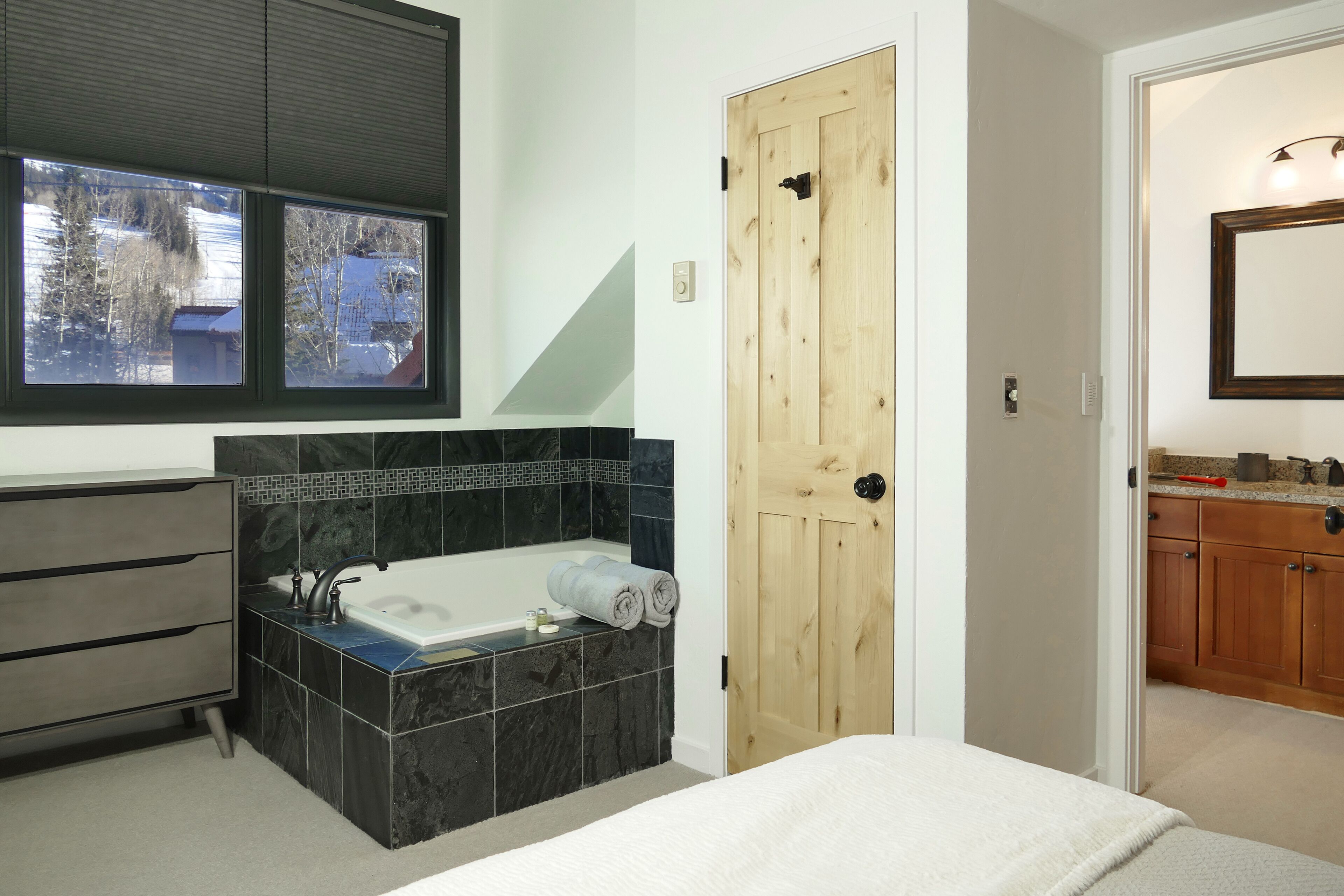 A modern bathroom with a bathtub, a wooden door, and a window with a view of trees and a building.