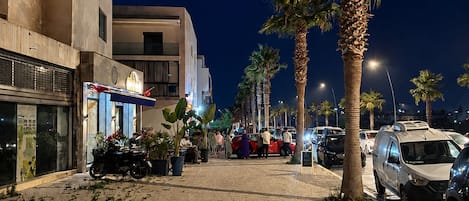 Una calle pavimentada bordeada de palmeras y coches aparcados, que conduce a un edificio con entrada visible.