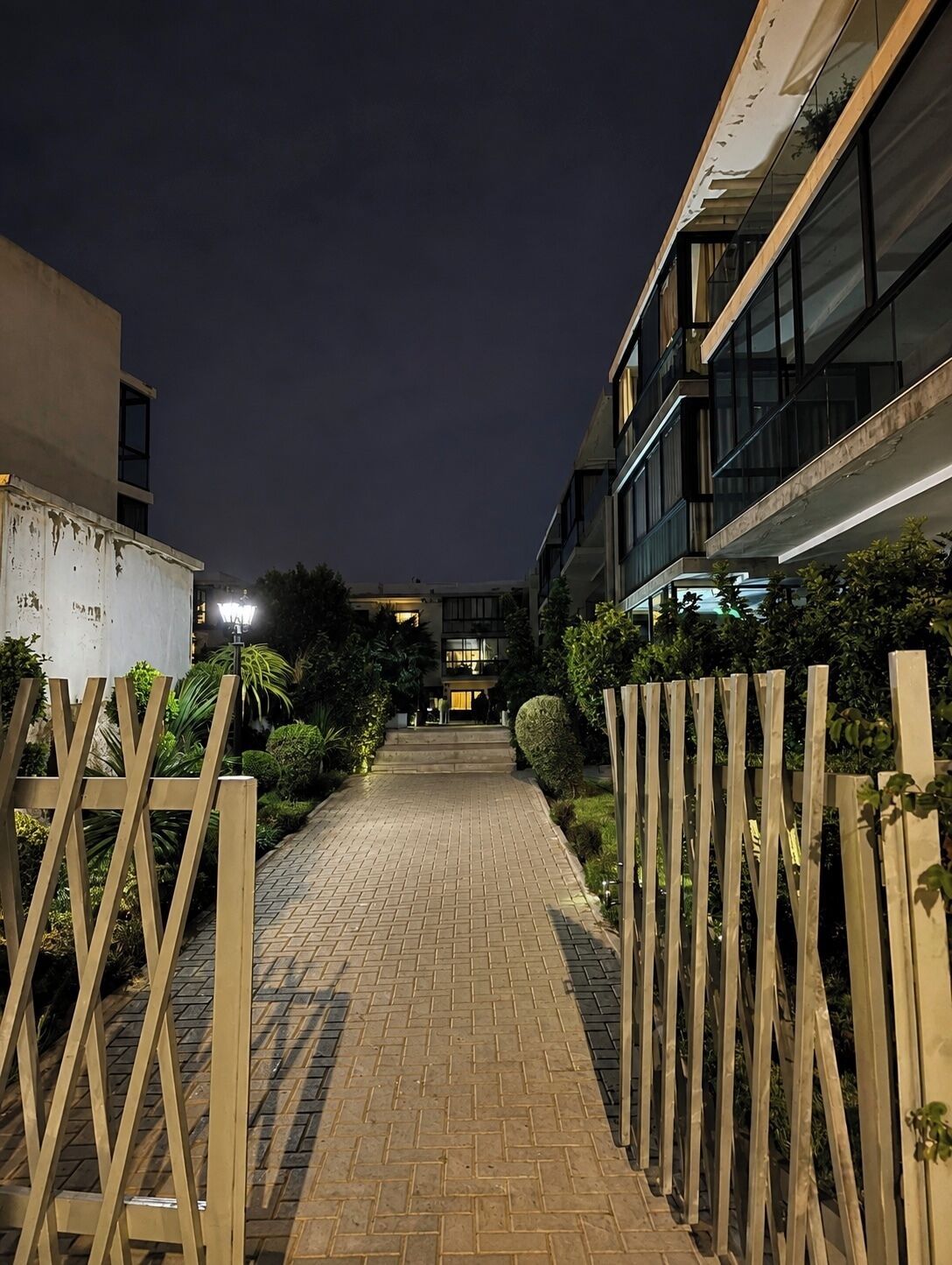 A pathway leading to a building with a modern design, surrounded by greenery and a wooden fence.