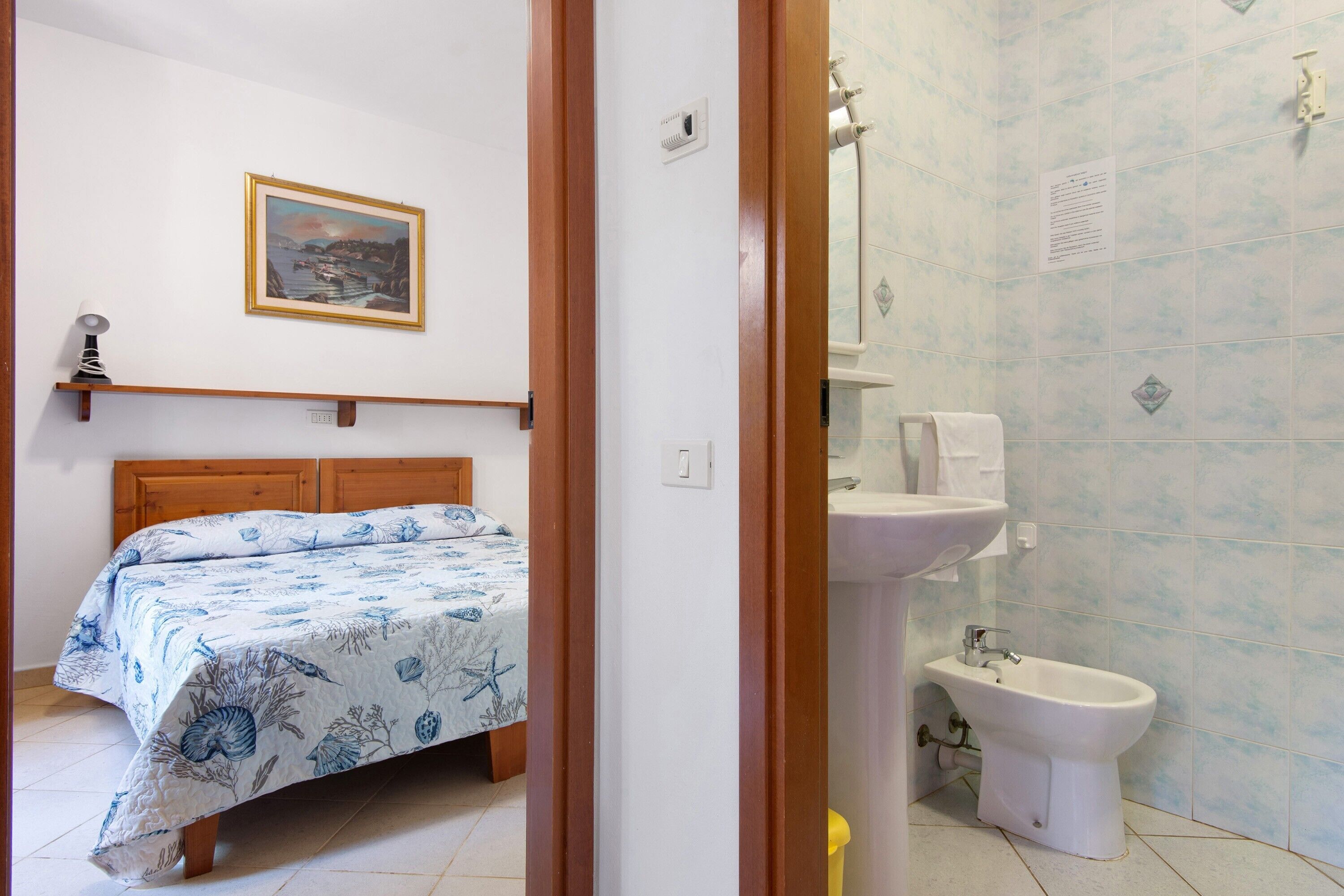 Ein Schlafzimmer mit Bett, Nachttischlampe, einem gerahmten Bild an der Wand und einem Badezimmer mit Waschbecken und Bidet.