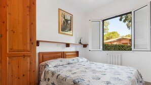 A bedroom with a wooden bed, a painting on the wall, and a window with a view of greenery. - Three-Room Apartment with Garden 100 Meters from the Beach - Sole e Mare Tourist Village (Rio)