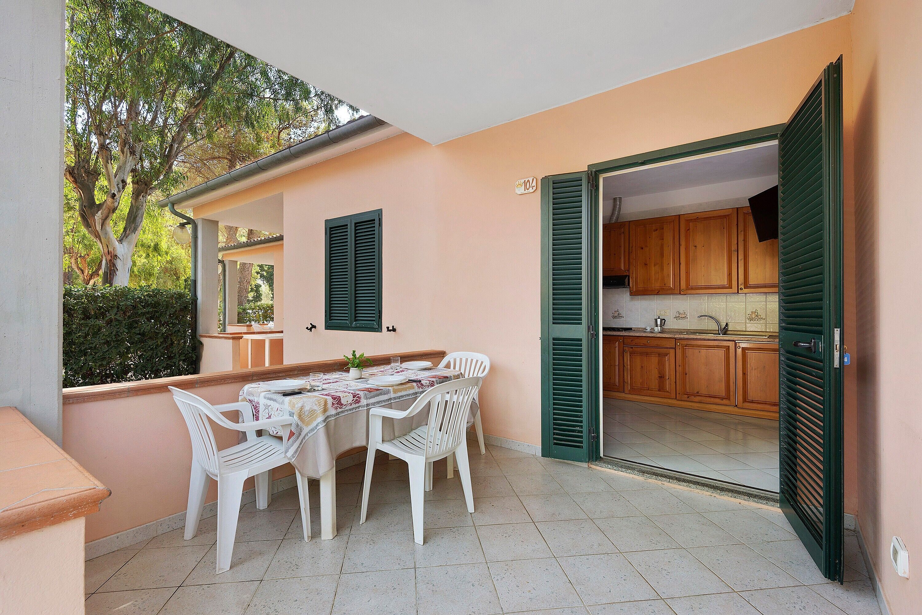 An outdoor dining area with a table and chairs, a view of the kitchen through an open door, and a tree in the background.