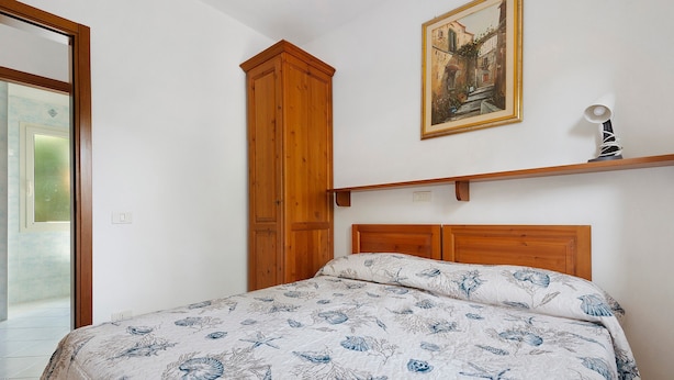 A bedroom with a bed, wooden wardrobe, a painting, and a shelf with a lamp. - Three-Room Apartment with Garden 100 Meters from the Beach - Sole e Mare Tourist Village (Rio)