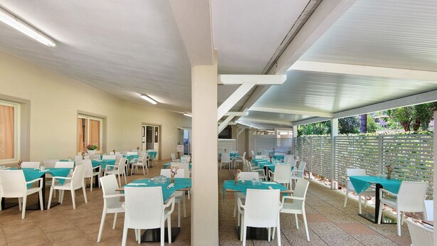 A restaurant with multiple tables and chairs, a tiled floor, and a ceiling with fluorescent lighting. - Three-Room Apartment with Garden 100 Meters from the Beach - Sole e Mare Tourist Village (Rio)