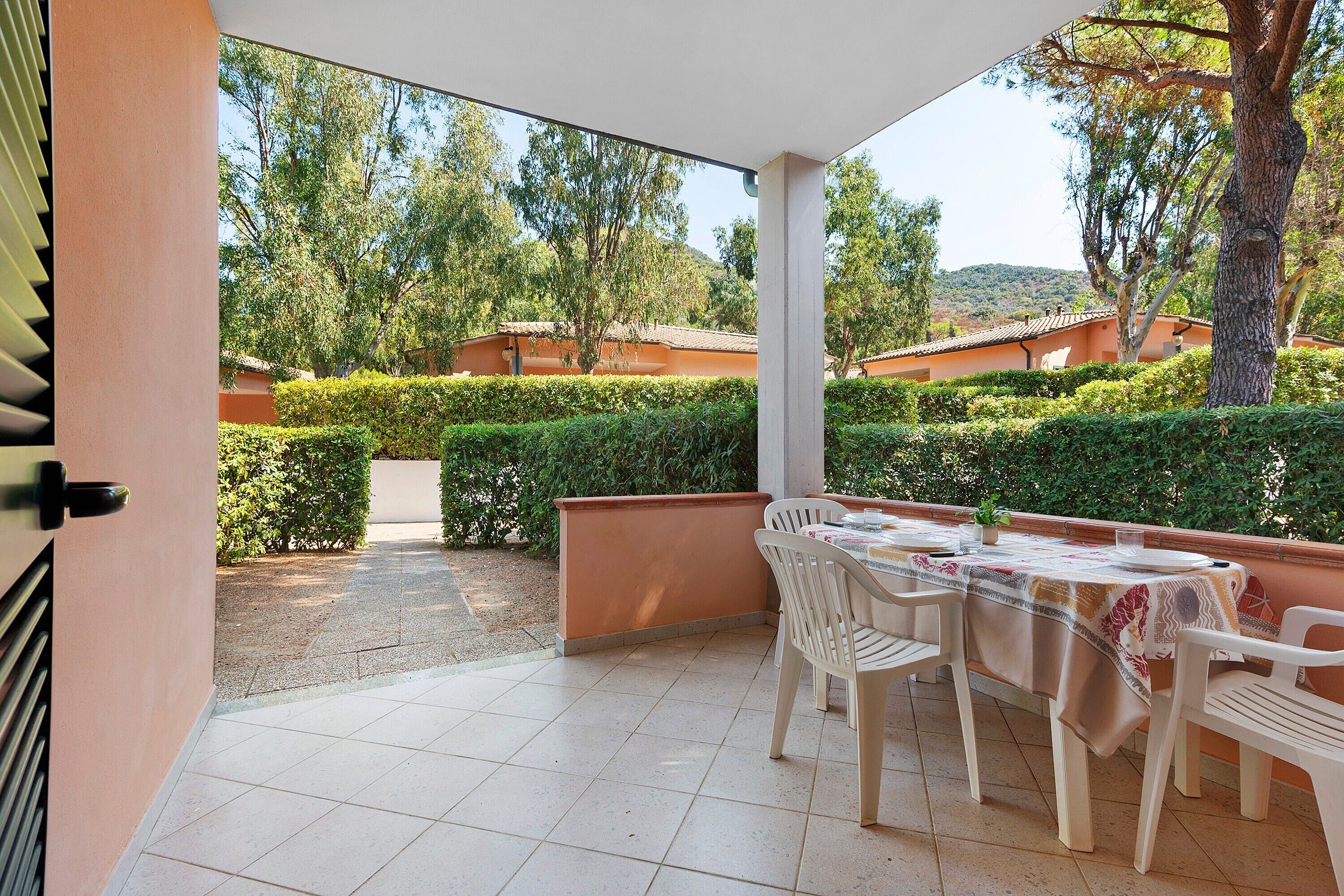 A covered patio with a table set for dining, surrounded by greenery and buildings in the background.