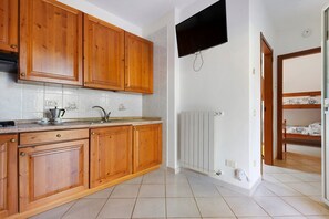 A kitchen with wooden cabinets, a built-in oven, a sink, and a wall-mounted television. - Three-Room Apartment with Garden 100 Meters from the Beach - Sole e Mare Tourist Village (Rio)