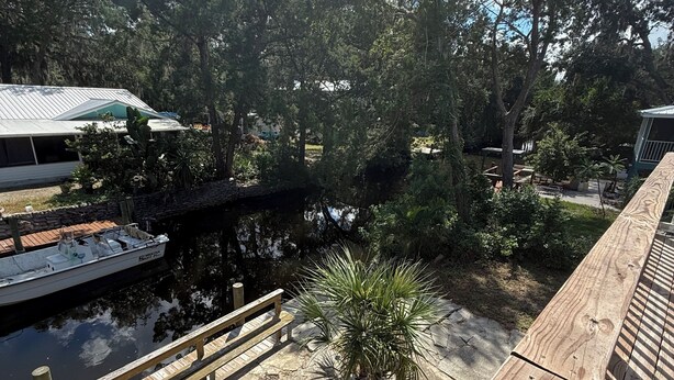 Una terraza de madera con una palmera pequeña, un muelle con un bote y un sendero de piedra.