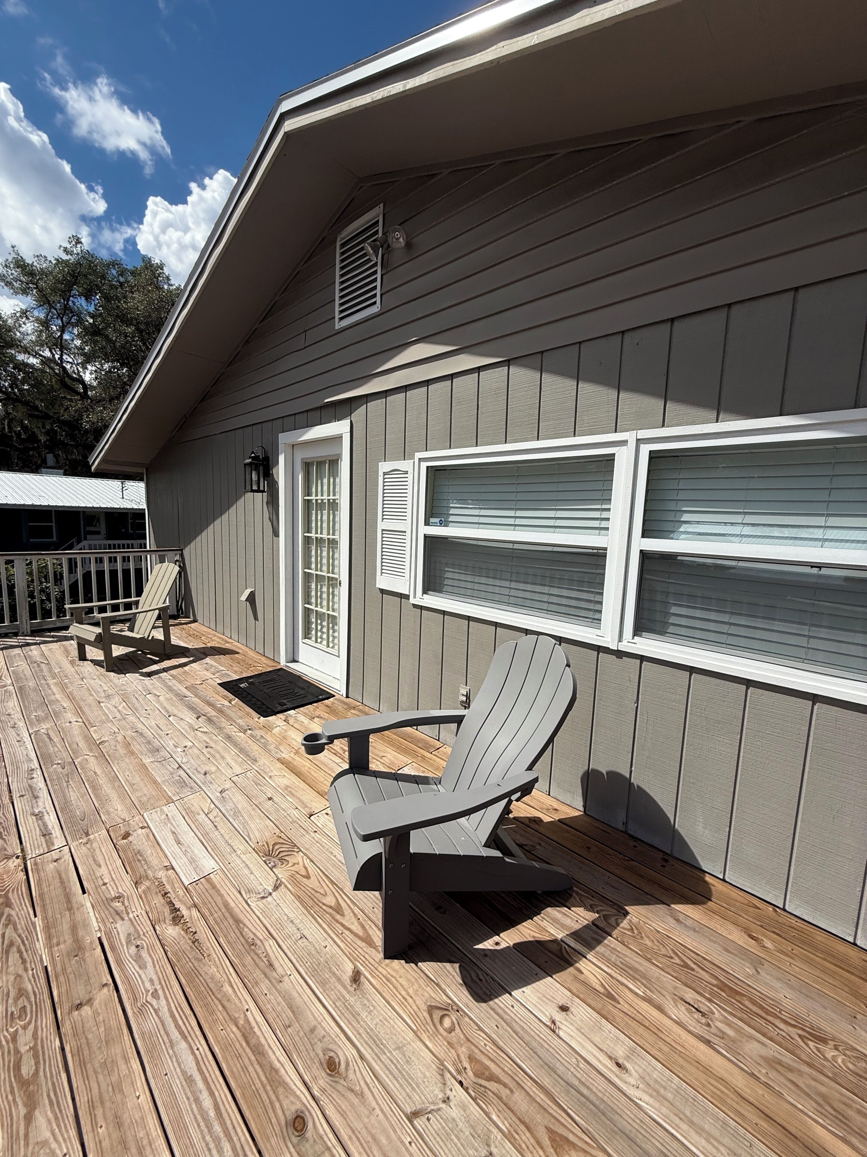 A wooden deck with Adirondack chairs and a covered patio area adjacent to a building with a window and a door.