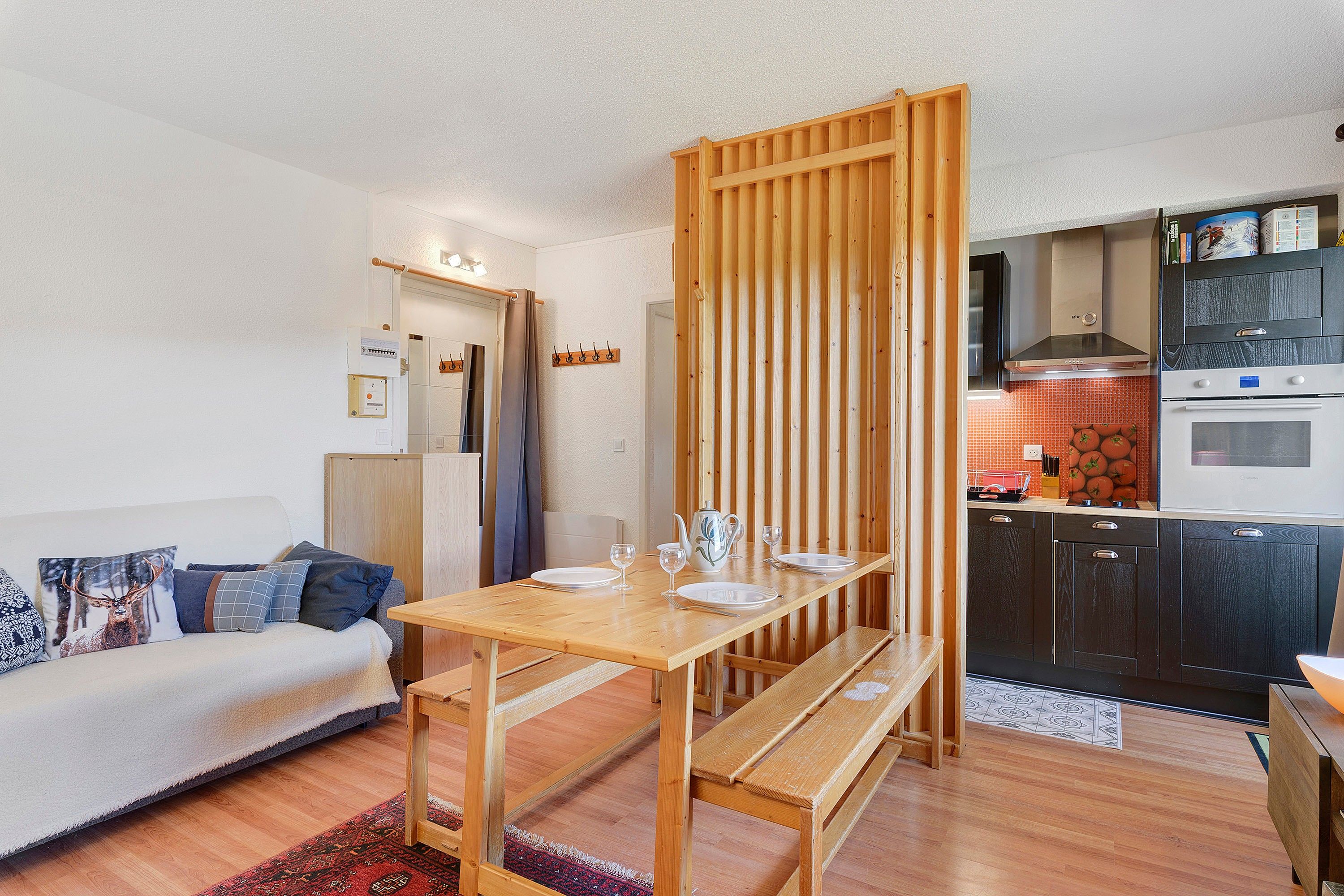 A compact living space with a kitchenette, a dining area with a wooden table and bench, and a sofa with patterned cushions.