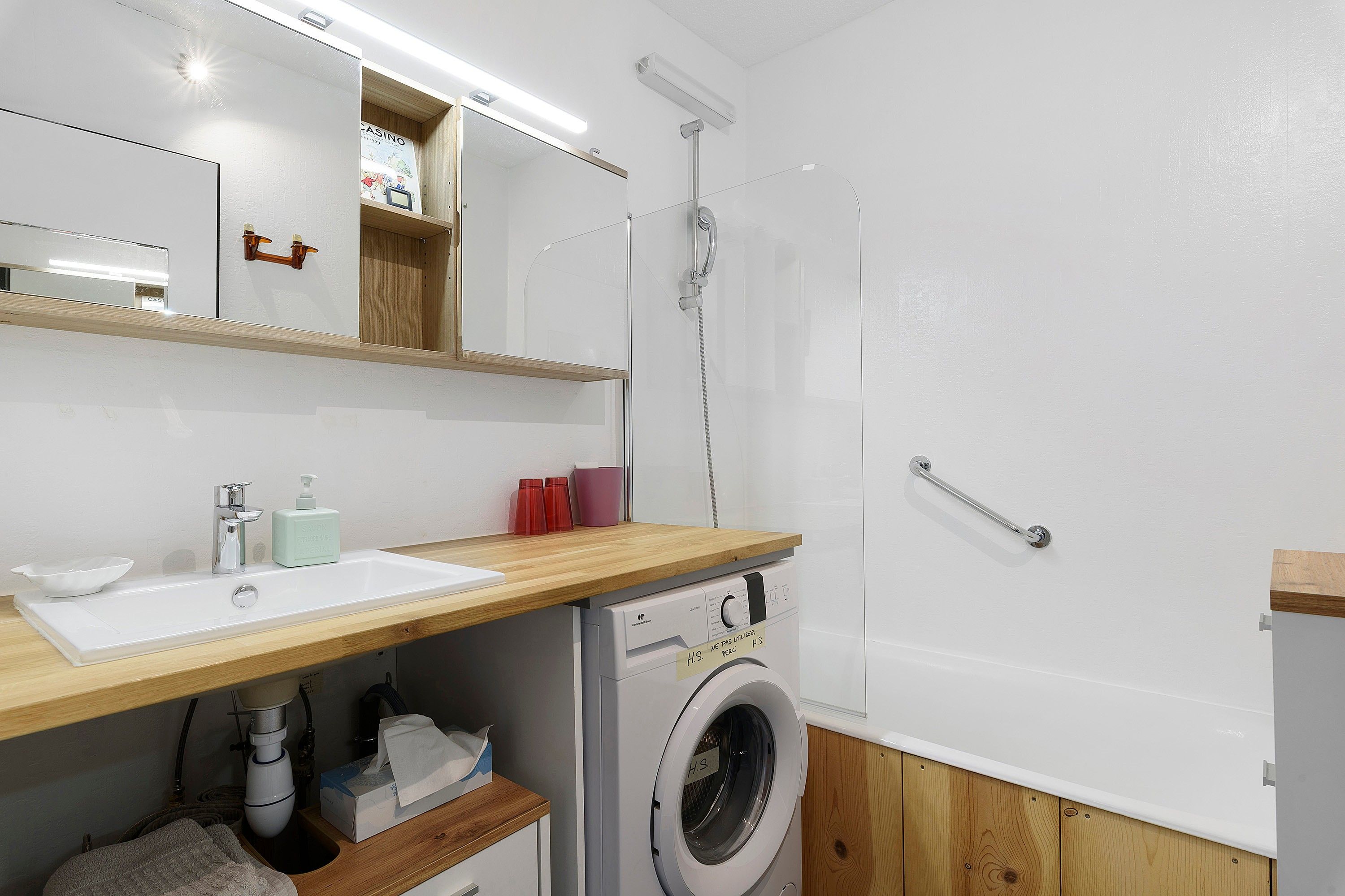 Une salle de bain moderne avec un meuble-lavabo en bois, un lave-linge encastré, une douche et un miroir avec un éclairage situé au-dessus.
