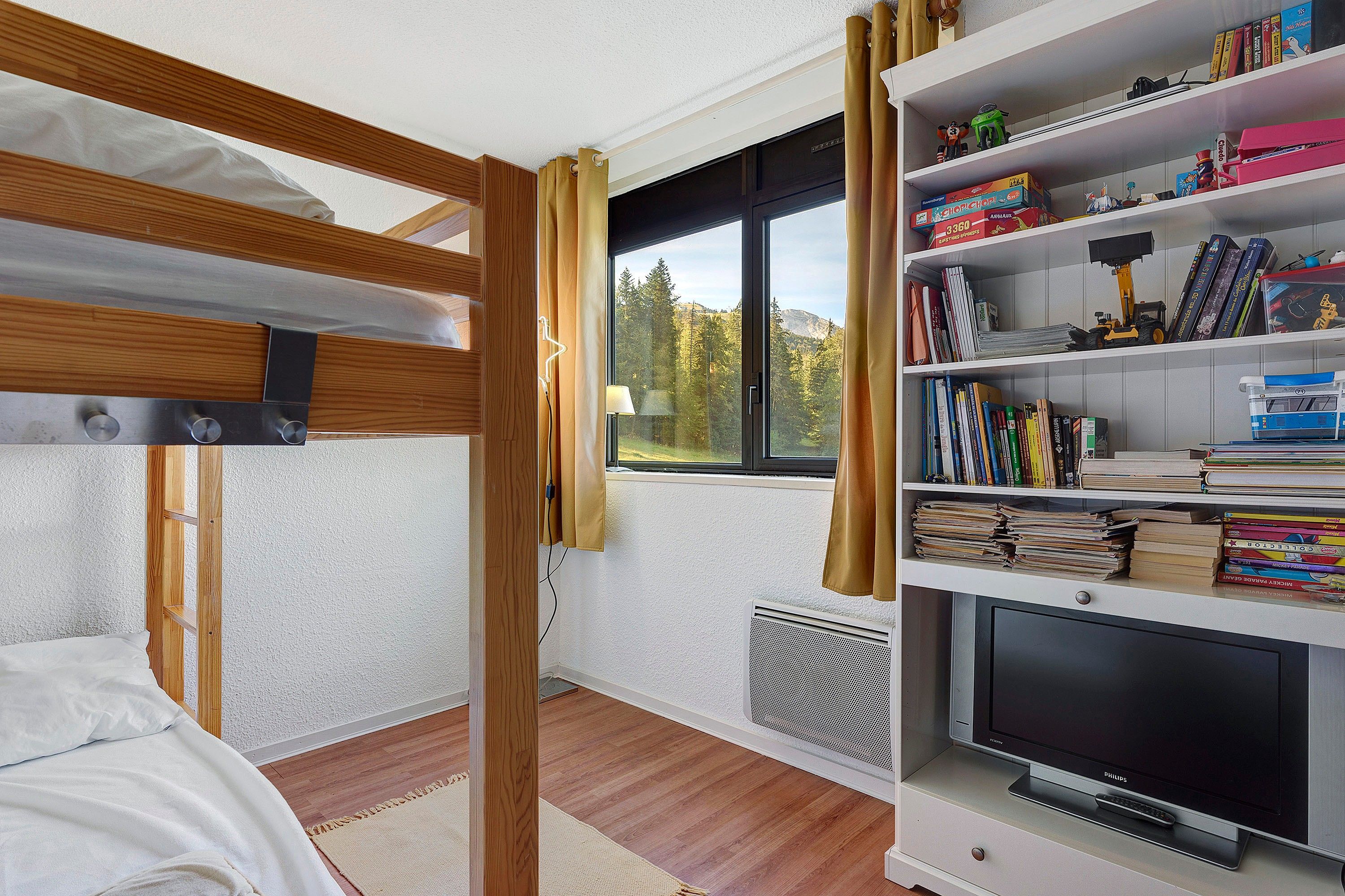Una habitación con literas, una estantería llena de libros y juguetes, un televisor y una ventana que da a árboles y montañas.