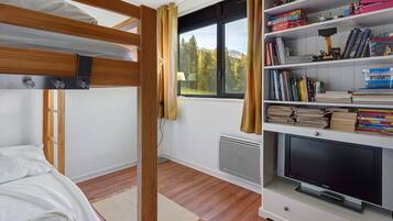 Una habitación con literas, una estantería llena de libros y juguetes, un televisor y una ventana que da a árboles y montañas.