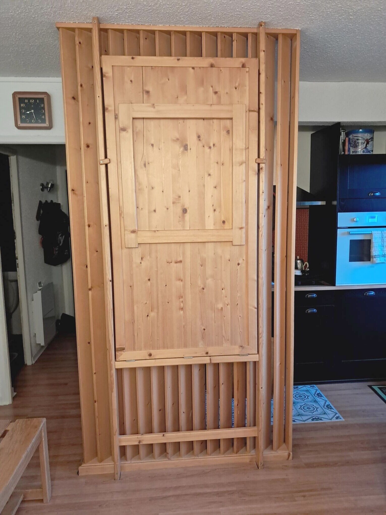 Una puerta corredera de madera con un sencillo sistema de cierre, un reloj en la pared y una cocina con un electrodoméstico azul.