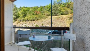 Un balcon avec une table et deux chaises, donnant sur un paysage naturel avec des arbres et une voiture au loin.