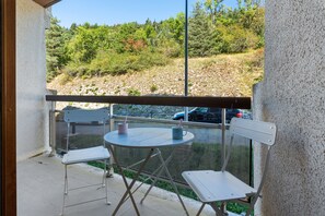 A balcony with a table and two chairs, overlooking a natural landscape with trees and a car in the background.