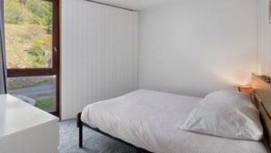 Une chambre à coucher avec un lit, une table de chevet, une fenêtre avec des stores et une vue sur la verdure à l’extérieur.