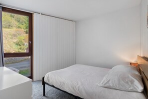 A bedroom with a bed, a bedside table, a window with blinds, and a view of greenery outside.